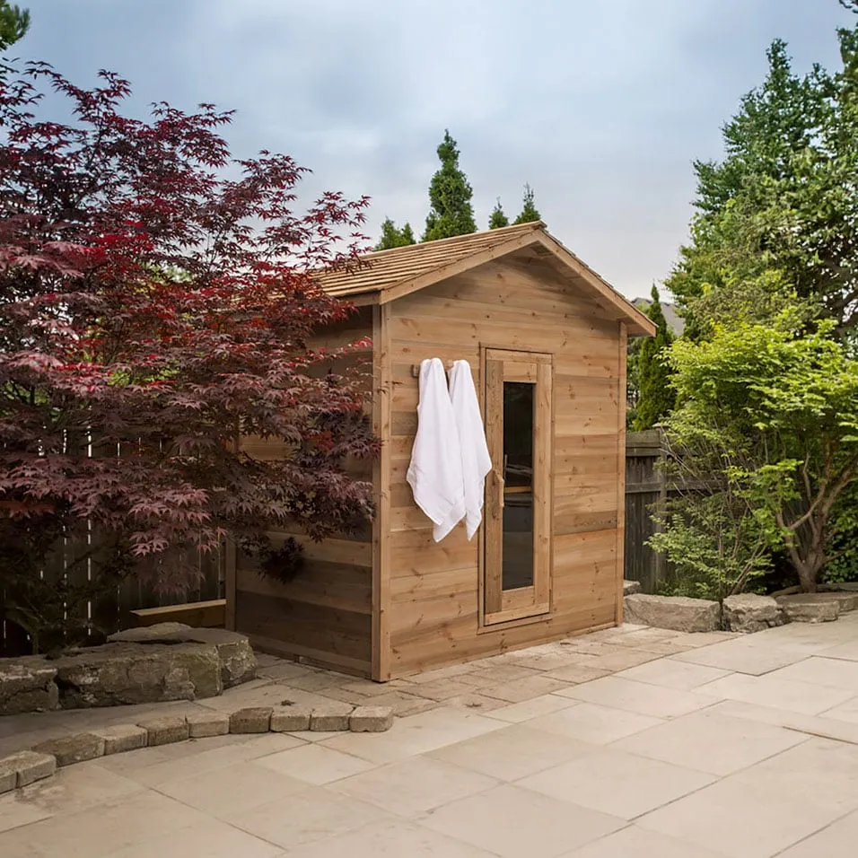 Cedar cabin sauna with gabled cedar shingle roof, glass-panel door and towels on a hook, on a stone patio beside a red maple
